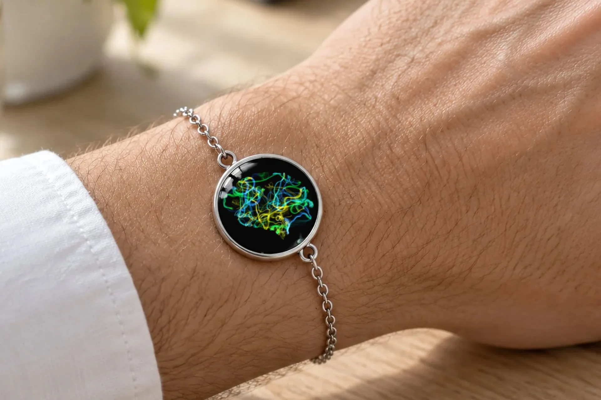 Un poignet posé sur une table en bois près d’une tasse de café : le bracelet CellandArt, une œuvre aux lignes vertes, jaunes et bleues sous un médaillon de verre.